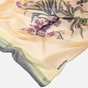 Detalle del tejido de seda y del estampado floral del pañuelo Surkana con flores en tonos morados y hojas en verde sobre fondo crema.