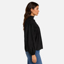 Vista lateral de una modelo con blusa negra Feeka de algodón 100%, cuello alto fruncido y mangas abullonadas.